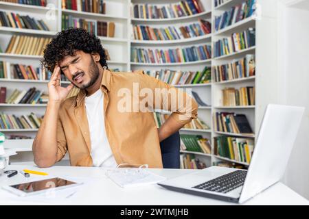 Homme hispanique malade ressentant des spasmes intenses dans la tête et le mal de dos tout en fermant les yeux dans la grimace de douleur. Freelance surmenée travaillant dans une position inconfortable et étant interrompue par un mauvais sentiment. Banque D'Images