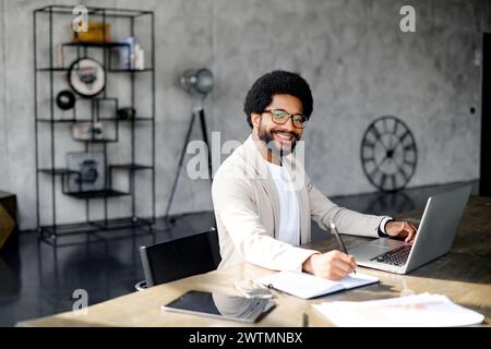 Un homme d'affaires brésilien joyeux en costume beige décontracté intelligent interagit avec son ordinateur portable dans un bureau moderne, son sourire transmettant satisfaction et positivité dans son travail Banque D'Images