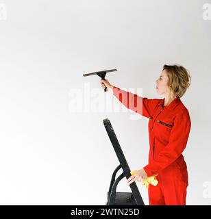 une jeune fille dans une combinaison rouge, une employée d'une entreprise de nettoyage, se tient sur un escabeau et tient un grattoir en verre dans sa main sur un b blanc isolé Banque D'Images