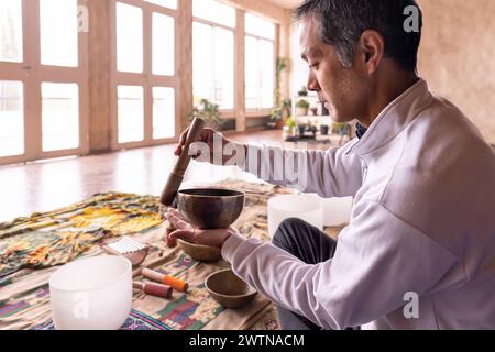 Un homme asiatique d'âge moyen est assis de profil, se concentrant sur la vibration douce d'un bol en laiton dans une pièce légèrement éclairée. Ancienne pratique de la méditation sonore dans Banque D'Images