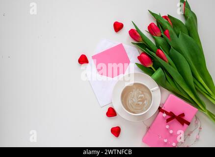 Bouquet de tulipes rouges, coffrets cadeaux avec des noeuds en ruban, tasse de café et carte de voeux sur la vue de dessus de table blanche. Banque D'Images