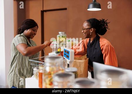 Cliente afro-américaine déchargeant son panier à la caisse. Vendeur de femme noire amicale avec un sourire, aidant l'acheteur avec ses produits alimentaires cultivés à la ferme. Banque D'Images