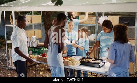 Fille caucasienne bénévole en distribuant de la nourriture gratuite aux nécessiteux lors d'un événement de collecte de nourriture à but non lucratif. Groupe de volontaires fournissant une aide humanitaire aux sans-abri et aux personnes en situation de pauvreté. Antenne, portable. Banque D'Images