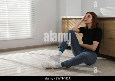 Triste jeune femme assise sur le sol dans la salle de bain, espace pour le texte Banque D'Images