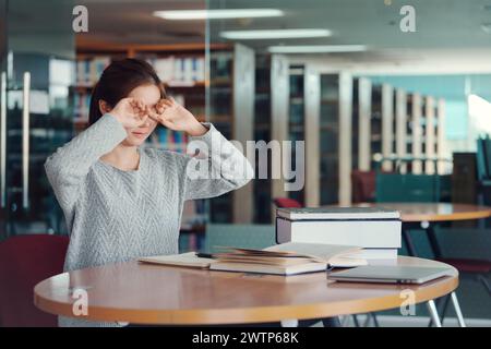 Jeune étudiante asiatique se frottant les yeux, se sentant fatiguée après avoir lu un livre à la bibliothèque. Banque D'Images