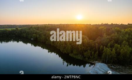 Cette photographie capture le moment serein d'un coucher de soleil au bord d'un lac tranquille niché au bord d'une forêt dense. Le soleil couchant jette une teinte chaude et dorée sur la scène, reflétant les eaux calmes du lac et soulignant la cime des arbres. La composition de l'eau, des bois et de la lueur du soleil crée un équilibre harmonieux entre les éléments, incarnant la splendeur tranquille de la nature. Lac serein à Forest's Edge pendant le coucher du soleil. Photo de haute qualité Banque D'Images
