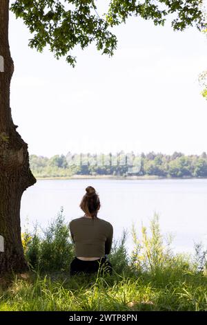 Une image sereine représentant une femme assise sous un arbre feuillu, regardant au-dessus d'un lac calme. La canopée naturelle au-dessus de la tête et l'étreinte douce de l'arbre créent une atmosphère de réflexion paisible. Femme embrassant la sérénité à Lakeside Under the Canopy. Photo de haute qualité Banque D'Images