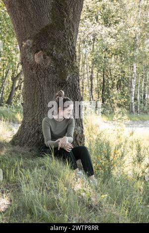 Cette image capture une jeune femme jouissant d'un moment de calme dans les bois, assise à la base d'un grand arbre avec la lumière du soleil filtrant à travers les feuilles, créant une atmosphère paisible et réfléchissante. Détendue jeune femme assise sous l'arbre dans Sun-Dappled Woods. Photo de haute qualité Banque D'Images