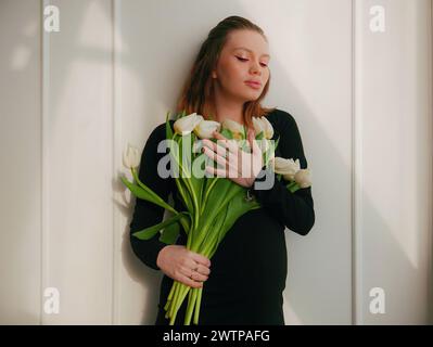 Une femme enceinte avec une coiffure et un maquillage sur son visage dans une robe serrée noire avec des tulipes blanches Banque D'Images