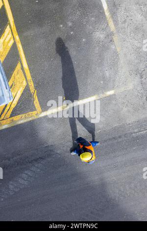 Un travailleur de la construction portant un casque de sécurité marche sur le tarmac. Il projette une longue ombre. Stuttgart, Bade-Wuerttemberg, Allemagne Banque D'Images