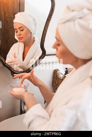 Portrait de femme heureuse appliquant la crème et les cosmétiques anti-âge sur le visage tout en regardant le miroir de réflexion portant en peignoir dans le spa après les procédures Banque D'Images