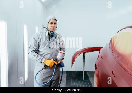 Main de peintre réparateur automobile dans un gant de protection avec pulvérisateur d'aérographe peignant la carrosserie de voiture dans la chambre de peinture. Banque D'Images