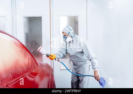 Gros plan d'un travailleur masculin en tissu de costume gris portant un masque de protection chimique à l'aide d'un pistolet pulvérisateur et d'un aérographe et peignant une voiture rouge. Banque D'Images