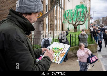 Le public vient admirer une nouvelle œuvre du mystérieux artiste Banksy sur la Hornsey Road au nord de Londres, le 18 mars 2024. La nouvelle pièce est apparue au cours du week-end, mais aujourd'hui l'artiste a revendiqué le crédit de cette dernière œuvre - un spray vert sur un mur résidentiel derrière un arbre à dos taillé pour ressembler à du feuillage, avec un pochoir d'une personne tenant un tuyau de pression à côté de lui. Banque D'Images