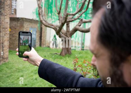 Le public vient admirer une nouvelle œuvre du mystérieux artiste Banksy sur la Hornsey Road au nord de Londres, le 18 mars 2024. La nouvelle pièce est apparue au cours du week-end, mais aujourd'hui l'artiste a revendiqué le crédit de cette dernière œuvre - un spray vert sur un mur résidentiel derrière un arbre à dos taillé pour ressembler à du feuillage, avec un pochoir d'une personne tenant un tuyau de pression à côté de lui. Banque D'Images