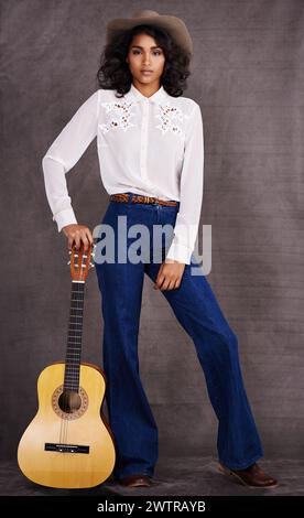 Femme, mode et musicienne cowgirl en studio, vêtements occidentaux et guitare avec confiance. Amérindien, personne féminine dans le portrait sérieux pour Banque D'Images