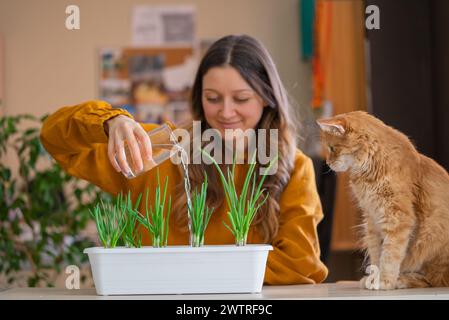 Une femme souriante dans un pull moutarde arde arrose des pousses d'oignons verts dans une jardinière tandis qu'un curieux chat au gingembre regarde, un moment de bonheur de jardinage à la maison Banque D'Images