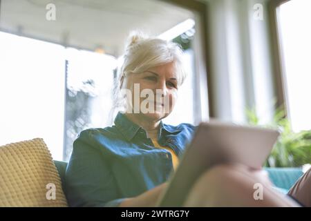 Femme mature utilisant la tablette numérique tout en étant assis sur le canapé à la maison Banque D'Images
