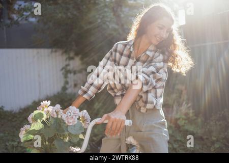 Une jeune femme attirante en vêtements décontractés est assise sur un vélo dans le jardin. Une charmante fille aux cheveux bouclés sourit dehors dans le soleil lumineux Banque D'Images