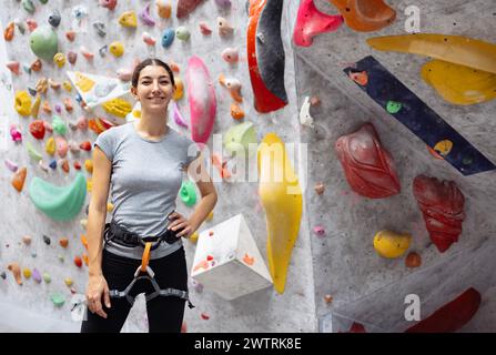 Jeune femme souriante en vêtements de sport posant près du mur d'entraînement. Une charmante brune grimpe sur un mur de blocs. Rire sport professionnel féminin cli Banque D'Images