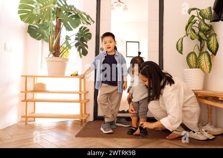 Une famille asiatique heureuse. Une jeune femme attirante aide à enlever les chaussures de sa petite fille. Une charmante mère coréenne et ses enfants sont revenus ho Banque D'Images