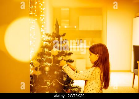 Une petite fille en pyjama décore un sapin de Noël dans la salle à manger. Un bébé mignon se prépare pour la nouvelle année. Un enfant heureux attend fo Banque D'Images