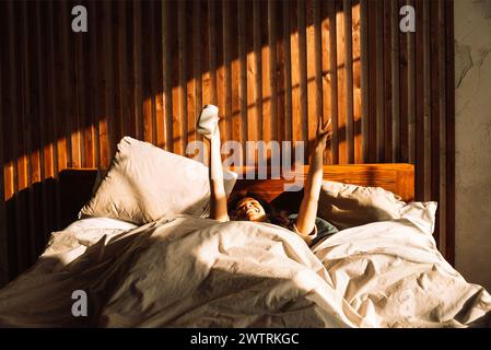 Charmant adolescent de race mixte rit et tient une tasse tout en étant allongé dans le lit. Mignonne fille africaine profite du matin ensoleillé. Charmant étudiant boit du café et sh Banque D'Images