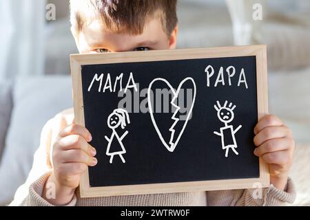 19 mars 2024 : petit garçon tenant une planche avec un dessin sur le divorce de ses parents. Image symbolique de la séparation et du divorce de maman et papa. PHOTOMONTAGE *** Kleiner Junge hält eine Tafel mit einer Zeichnung über die Scheidung der Eltern. Symbolbild Trennung und Scheidung von Mama und Papa. FOTOMONTAGE Banque D'Images