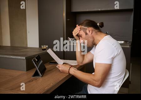 Jeune homme assis à la table, mettant sa main sur son front, ressentant frustration et choc par les coûts de paiement des factures Banque D'Images