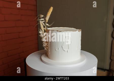 Gâteau blanc festif d'anniversaire à un étage dans un style rustique, épillets de blé sec décorés et glaçage à la crème vanille sur le stand du restaurant de la réception. Mariage, fête d'anniversaire. Service traiteur en événement. Banque D'Images