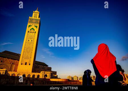 Une personne portant un foulard rouge vif regarde une tour de mosquée majestueuse contre un ciel bleu profond au coucher du soleil. Banque D'Images