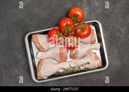 Cuisses de poulet crues dans une palette en métal. Branche verte avec tomates. Champignons dans un bol. Fond noir. Pose à plat Banque D'Images