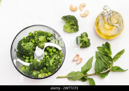 Pois verts, brocoli et noix de cajou dans un bol mélangeur. Salade romaine et menthe, bouteille d'huile d'olive sur table. Pose à plat. Fond blanc. Banque D'Images