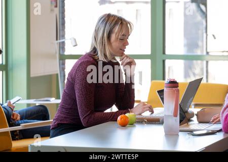 Une femme concentrée travaillant sur son ordinateur portable tout en profitant d'une collation saine dans un espace de coworking lumineux. Banque D'Images