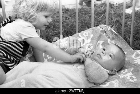 Années 1970, historique, dans un parc en bois, une petite fille interagissant avec son frère bébé allongé sur le dos regardant vers le haut, Angleterre, Royaume-Uni. Banque D'Images