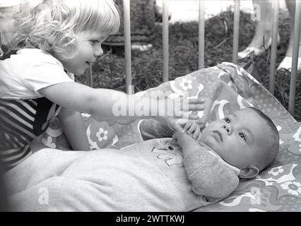 Années 1970, historique, une petite fille interagissant avec son petit frère allongé sur le dos iin un parc en bois, Angleterre, Royaume-Uni. Banque D'Images