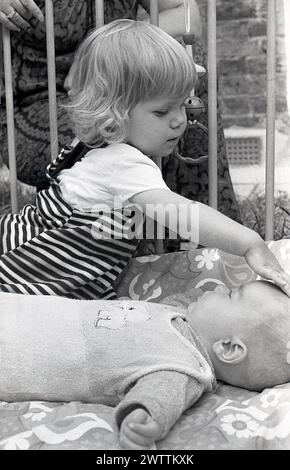 Années 1970, historique, une petite fille interagissant avec son petit frère allongé sur le dos iin un parc en bois, Angleterre, Royaume-Uni. Banque D'Images