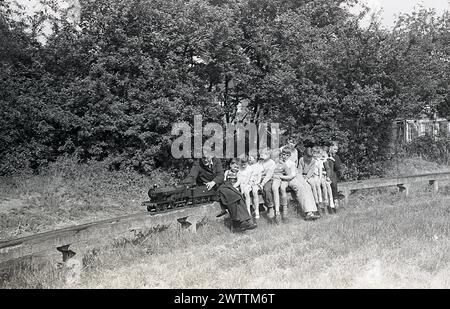 Années 1960, historique, été et les enfants appréciant une balade sur un chemin de fer miniature ou jardin, avec un conducteur de train en uniforme avec chapeau conduisant le train à vapeur miniature sur la piste elavated, Angleterre, Royaume-Uni. Banque D'Images