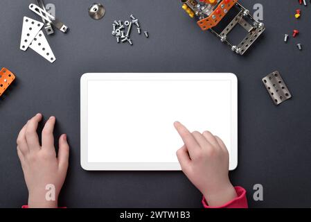 Maquette de tablette sur le bureau. Les mains de l'enfant touchent l'écran isolé. Vue de dessus, composition de bureau plat avec petites pièces métalliques et vis Banque D'Images