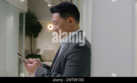 Asiatique d'âge moyen souriant homme d'affaires chinois japonais coréen heureux homme d'affaires employeur PDG directeur général près du mur dans la navigation dans le hall de bureau Banque D'Images