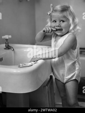 Une jeune fille se tient devant un évier de salle de bain, se brossant les dents avec du dentifrice tout en regardant la caméra. Elle porte une couche et un noeud dans les cheveux. Banque D'Images