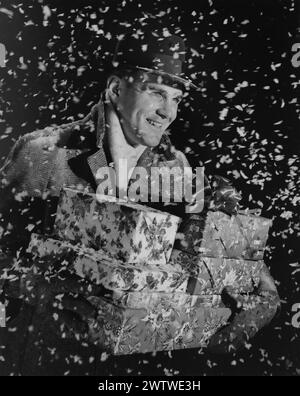 Homme d'âge mûr photographié comme de la fausse neige tourbillonne autour de lui. Il porte un chapeau, un manteau épais et des gants portant une pile de cadeaux de Noël Banque D'Images