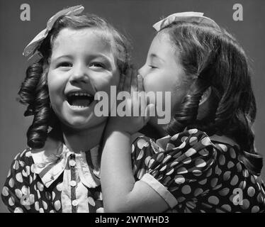 Deux jeunes filles jumelles habillées pareillement avec des robes polk-a-dot et un noeud dans leurs cheveux. Une fille murmure dans l'oreille de l'autre fille tandis que l'autre rit Banque D'Images