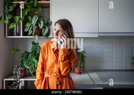 Femme fleuriste d'âge moyen parlant sur le téléphone portable, regardant la fenêtre debout dans la cuisine de la maison. Banque D'Images