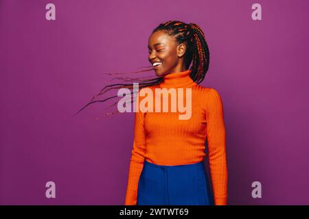 Femme africaine Gen Z avec des tresses élégantes à deux tons se tient dans un studio avec un fond violet vibrant. Elle porte des vêtements décontractés, y compris des jeans et un Banque D'Images