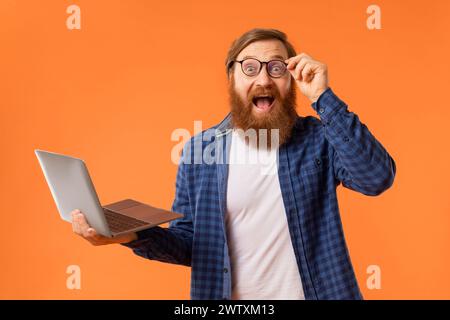 Un programmeur barbu aux cheveux roux excité tient un ordinateur portable sur fond orange Banque D'Images