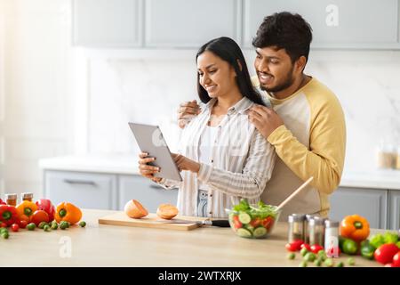 Heureux aimant jeunes conjoints indiens utilisant la tablette numérique pendant la cuisson Banque D'Images