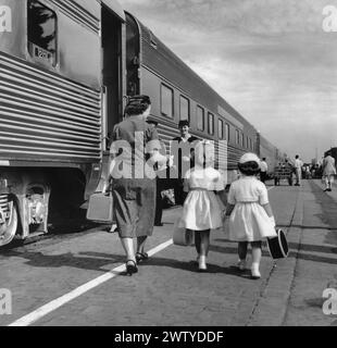 Mère avec ses deux petites filles en robes blanches marchant à côté d'un train de passagers se préparant à embarquer Banque D'Images