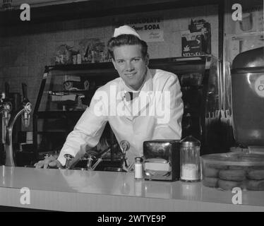 Jeune homme vêtu d'un manteau blanc, d'un noeud papillon et d'un chapeau blanc, souriant à la caméra tout en se tenant derrière le comptoir d'un magasin de soda Banque D'Images