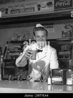 Jeune homme vêtu d'un manteau blanc, d'un noeud papillon et d'un chapeau blanc, souriant à la caméra tout en se tenant debout derrière le comptoir d'un magasin de boissons gazeuses offrant un milkshake et un sourire. Banque D'Images